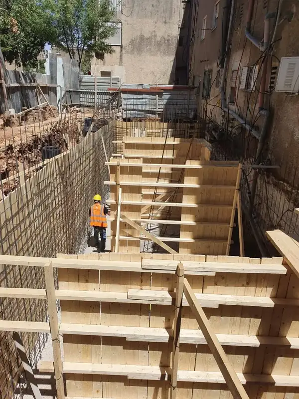 איש צוות בקסדה ואפוד זוהר בתוך חפירת המרתף, בין תבניות היציקה לקיר הבניין הקיים